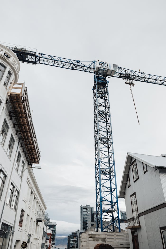 Tower crane in urban setting with buildings under cloudy sky, vertical shot.